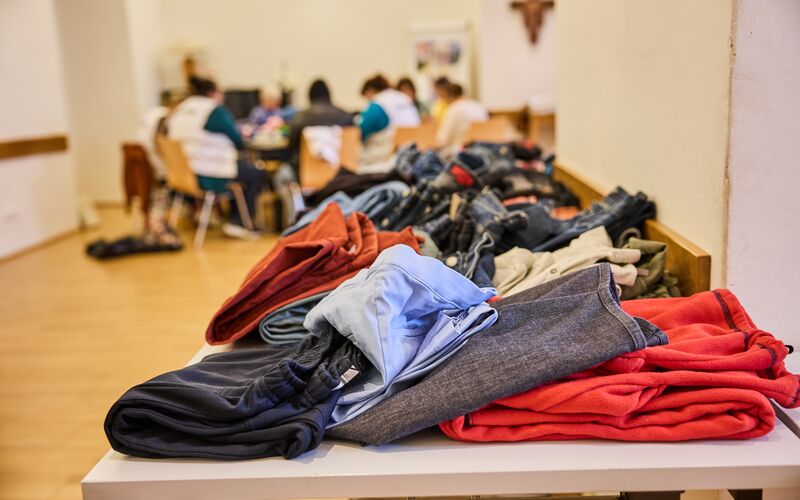 Ein Tisch mit gefalteter Kleidung, darunter Hosen und Pullover, während Menschen im Hintergrund sich unterhalten.