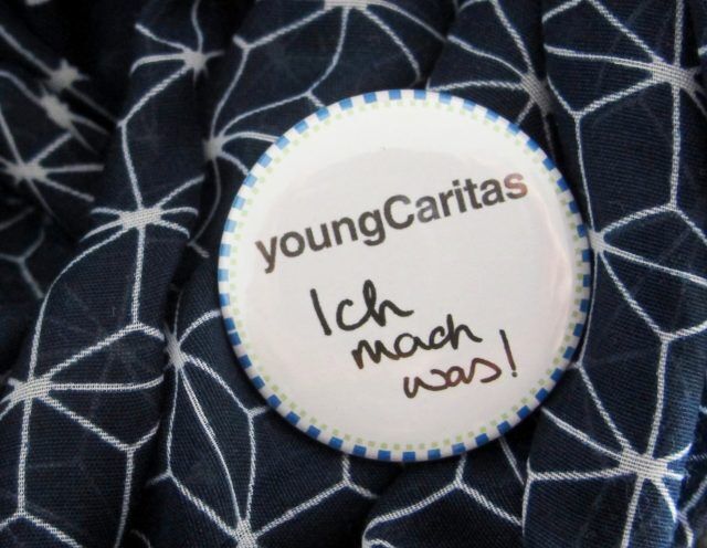 Ein Anstecker mit dem Schriftzug 'youngCaritas' und der handschriftlichen Aufschrift 'Ich mach was!'. Er liegt auf einem Stoff mit geometrischem Muster.