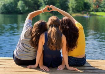 Drei Frauen sitzen auf einem Holzsteg am Wasser und bilden mit ihren Händen ein Herz.