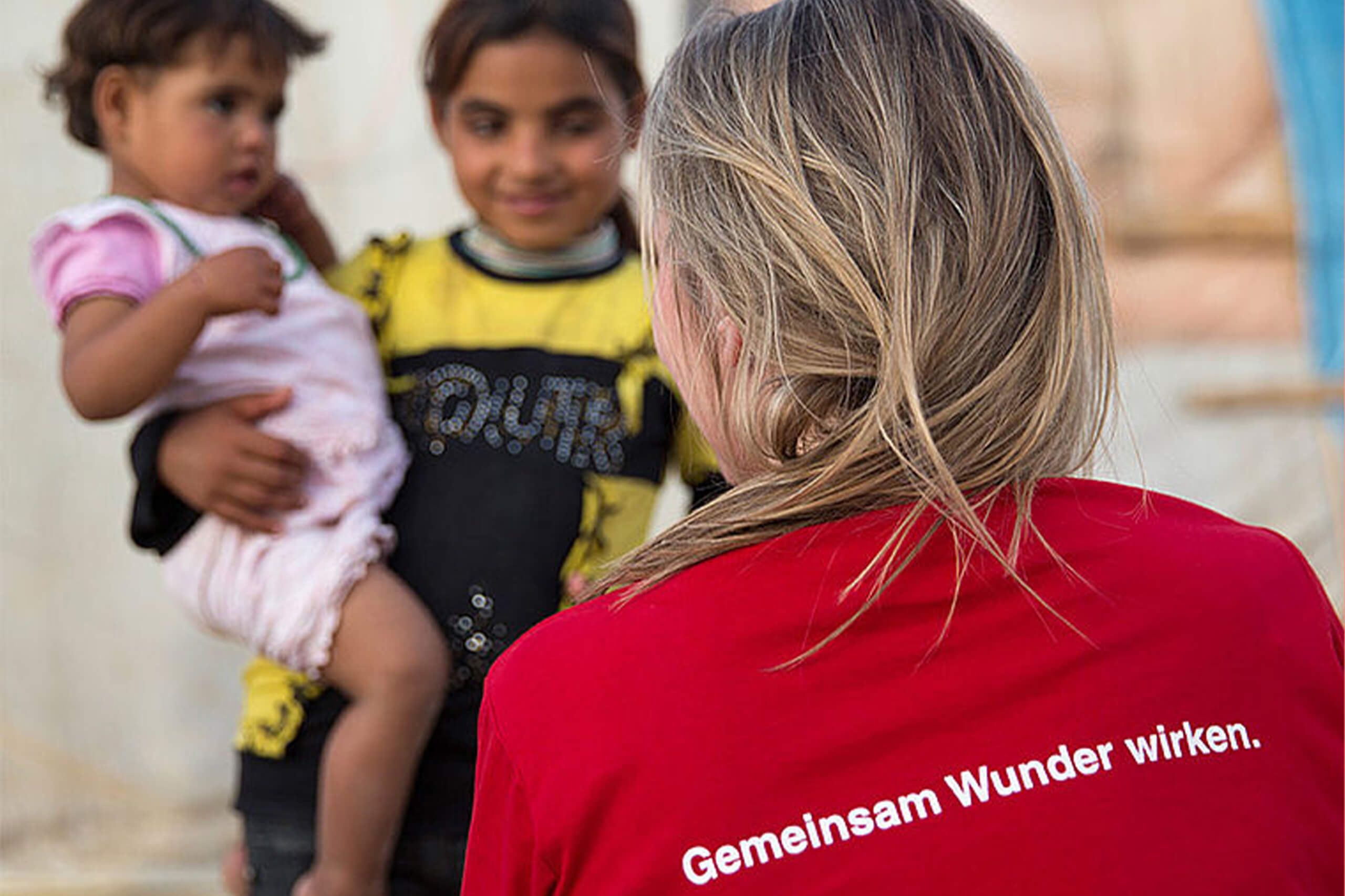 Eine Person im roten Shirt, die mit Kindern in einem Lager spricht. Die Botschaft ist, gemeinsam gute Taten zu vollbringen.
