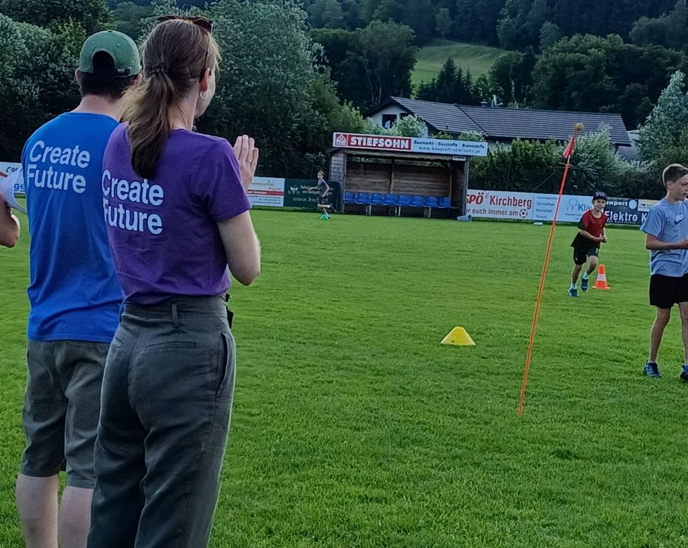 Zwei Personen in T-Shirts mit "Create Future" beobachten ein sportliches Training auf einem grünen Feld, während Kinder aktiv spielen.