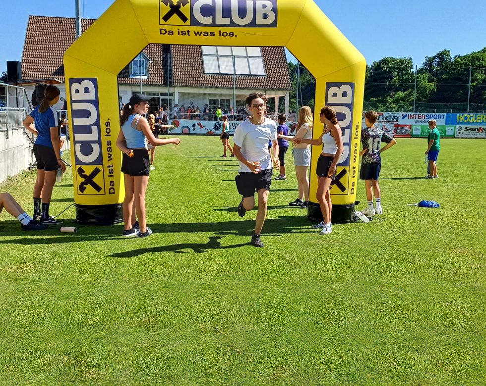 Junge Leute beim Sportfest, laufen unter einem bunten Zielbogen vorbei, viele Zuschauer im Hintergrund.