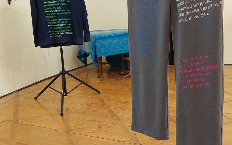 Zwei Kleidungsstücke auf Ständern in einem Ausstellungsraum, mit Botschaften über Umweltbewusstsein und Modekonsum.