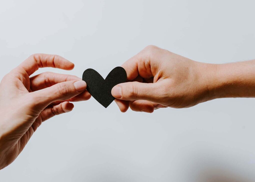Zwei Hände halten ein schwarzes Papierherz, symbolisieren Verbindung und Liebe.