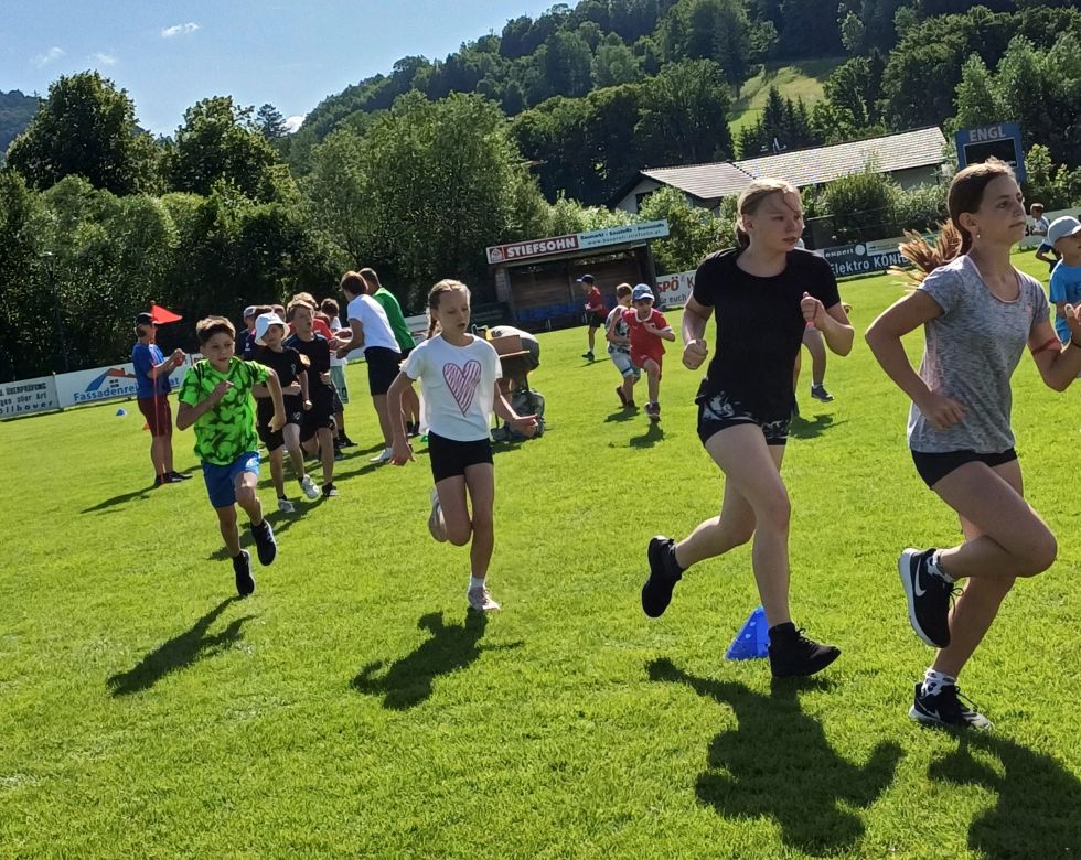 Eine Gruppe von Kindern läuft auf einem sonnigen Feld. Verschiedene Trikots und Shorts sind zu sehen, samt Fähnchen und Trainingseinrichtungen.