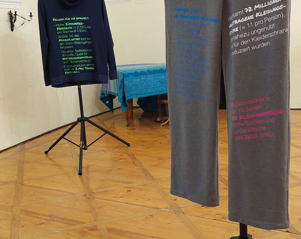 Zwei Kleidungsstücke auf Ständern in einem Ausstellungsraum, mit Botschaften über Umweltbewusstsein und Modekonsum.