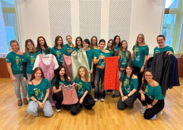 Eine Gruppe von Frauen steht lächelnd zusammen und hält verschiedene Kleider in den Händen, während sie bunte T-Shirts tragen.