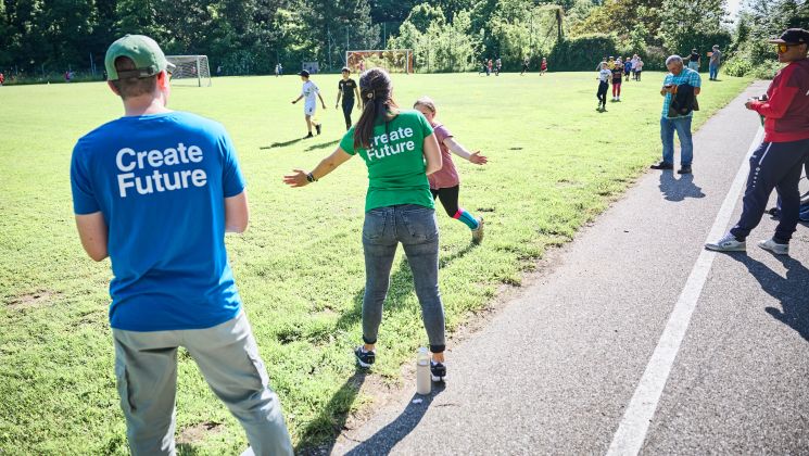 Eine Gruppe von Kindern spielt auf einem Fußballplatz, während Erwachsene zuschauen. Ein Mädchen rennt zu einer Frau in einem grünen Shirt.