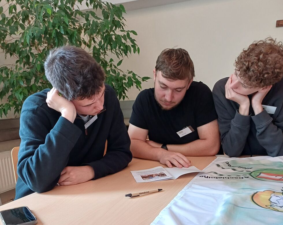 Drei junge Männer sitzen an einem Tisch und lesen konzentriert in einem Heft während einer besinnlichen Diskussion.