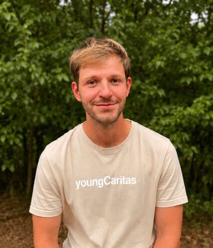 Ein junger Mann trägt ein cremefarbiges T-Shirt mit der Aufschrift youngCaritas und lächelt.