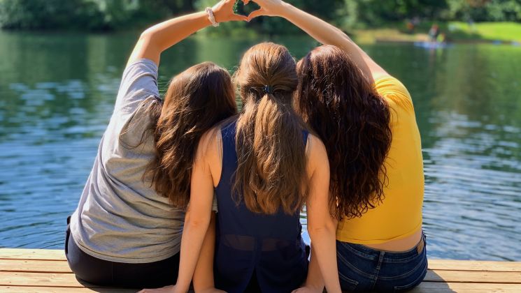 Drei Frauen sitzen auf einem Holzsteg am Wasser und bilden mit ihren Händen ein Herz.