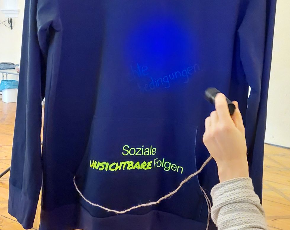 Ein schwarzer Hoodie, auf den mit einem Laserpointer Worte projiziert werden, darunter "Soziale Unsichtbare Folgen".