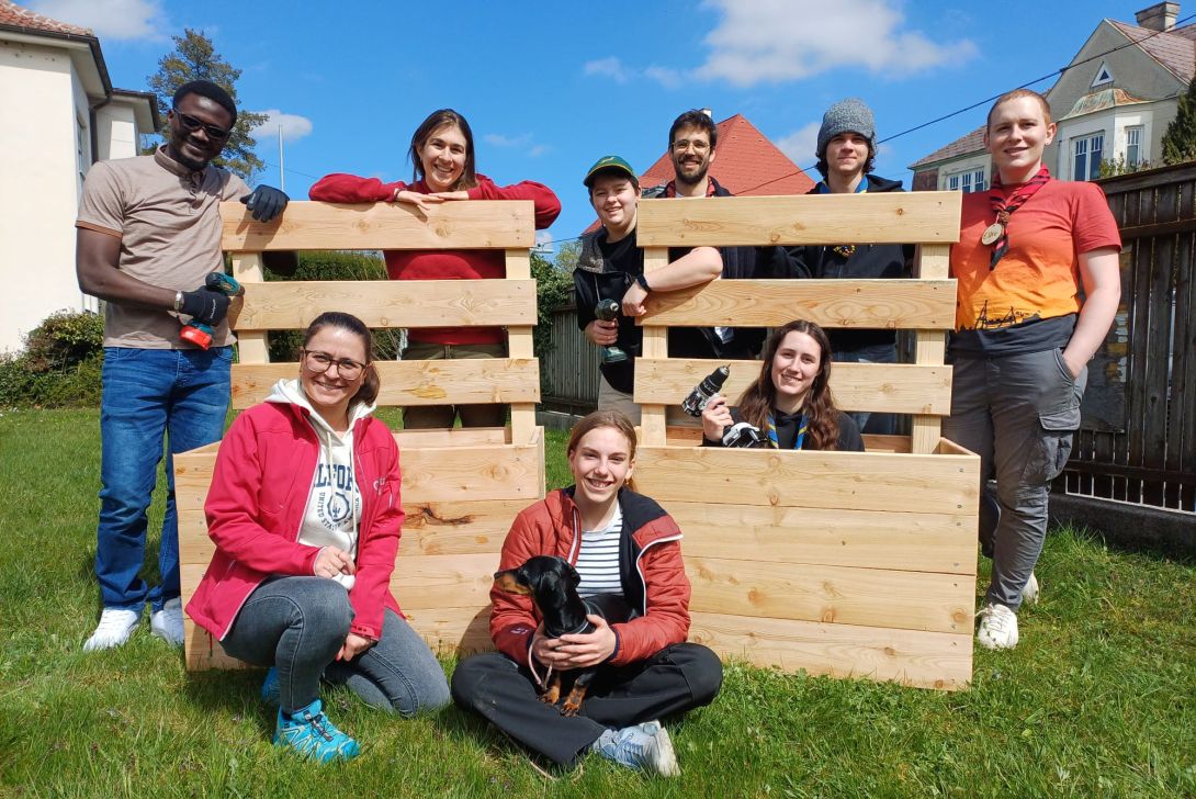 Eine Gruppe von Freiwilligen lächelt vor Holzpaletten im Freien, während sie an einem Projekt arbeiten.