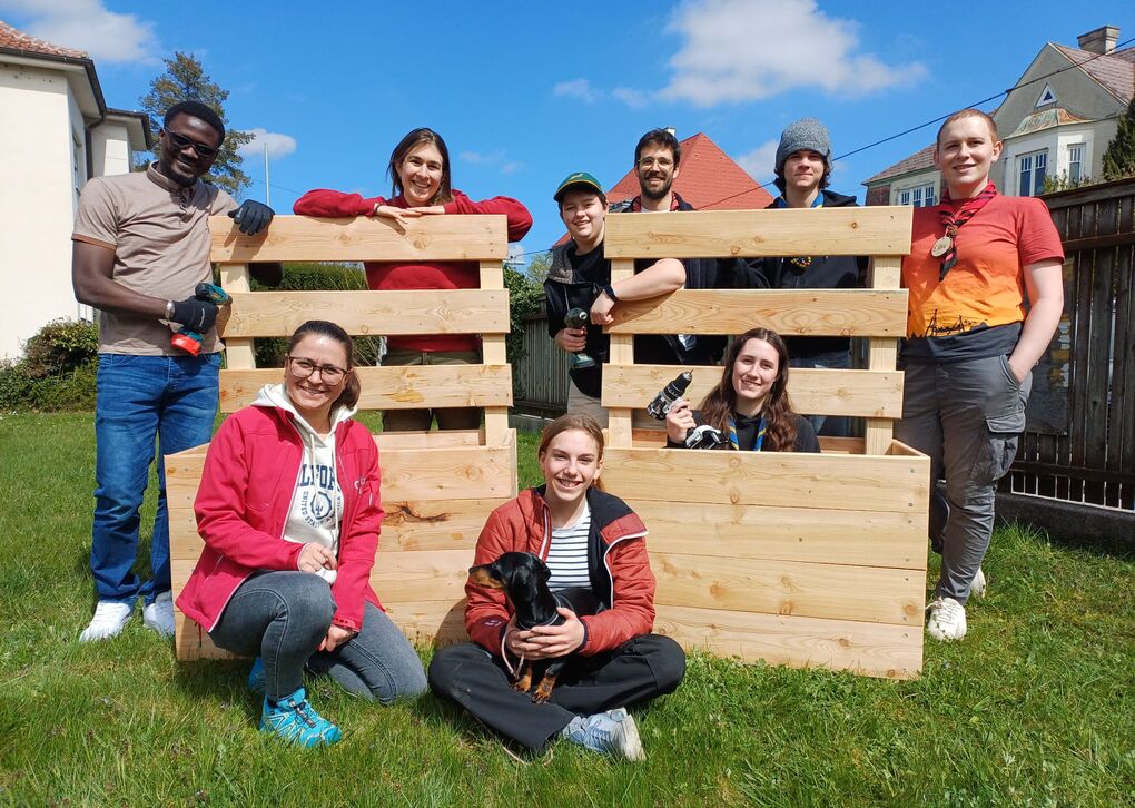Menschen stehen rund um ein aus Holz gebautes Beet im Freien, umgeben von einer Wiese und Himmel.