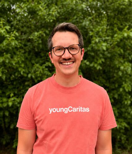 Ein junger Mann trägt ein lachsfarbenes T-Shirt mit der Aufschrift youngCaritas und lächelt