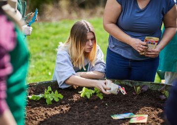 Eine Gruppe von Menschen, die Samen in einem Garten pflanzen, während eine Person konzentriert arbeitet und anderen zuschaut.