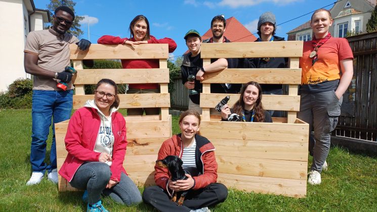 Eine Gruppe von Freiwilligen lächelt vor Holzpaletten im Freien, während sie an einem Projekt arbeiten.