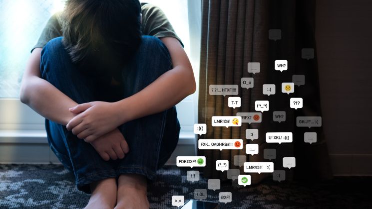Ein Junge sitzt auf dem Boden, umgeben von negativen Nachrichten und Emojis auf dem Bildschirm, die seine Traurigkeit und Isolation widerspiegeln.