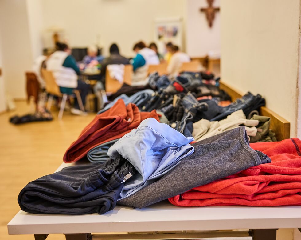 Ein Tisch mit gefalteter Kleidung, darunter Hosen und Pullover, während Menschen im Hintergrund sich unterhalten.