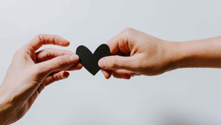 Zwei Hände halten ein schwarzes Papierherz, symbolisieren Verbindung und Liebe.