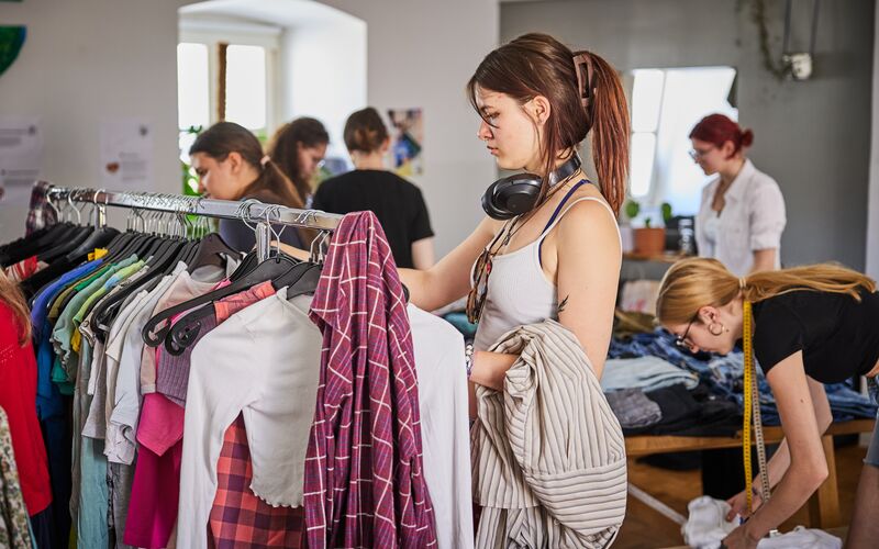 Eine Gruppe von Menschen durchstöbert Kleidung bei einer Second-Hand-Veranstaltung. Eine Frau wählt Einzelteile aus einem Kleiderständer aus.