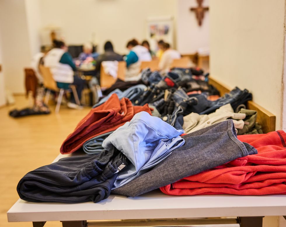Ein Tisch mit gefalteter Kleidung, darunter Hosen und Pullover, während Menschen im Hintergrund sich unterhalten.