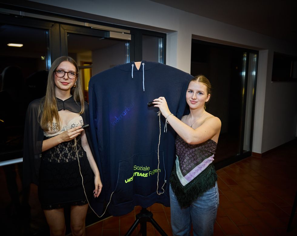 Zwei Frauen präsentieren einen Pullover mit verschiedenen Aufschriften während einer Veranstaltung.