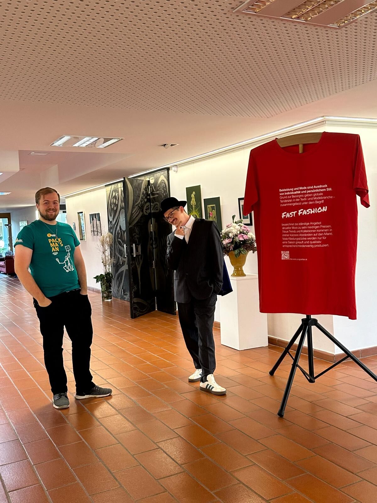 Zwei junge Männer posieren neben einem großen roten T-Shirt, das auf das Thema Fast Fashion aufmerksam macht.