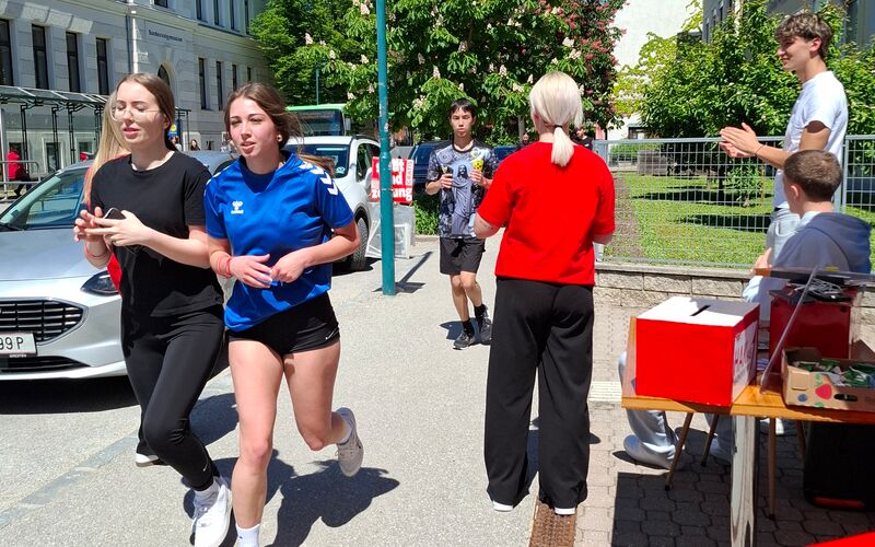 Schüler*innen laufen am Gehsteig an einem Stand vorbei und werden angefeuert.