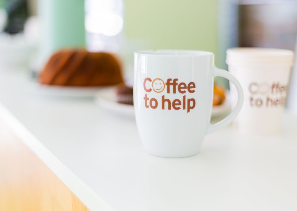 Eine weiße Kaffeetasse mit der Aufschrift 'Coffee to help' steht auf einem Tisch, umgeben von Kuchen und Keksen.