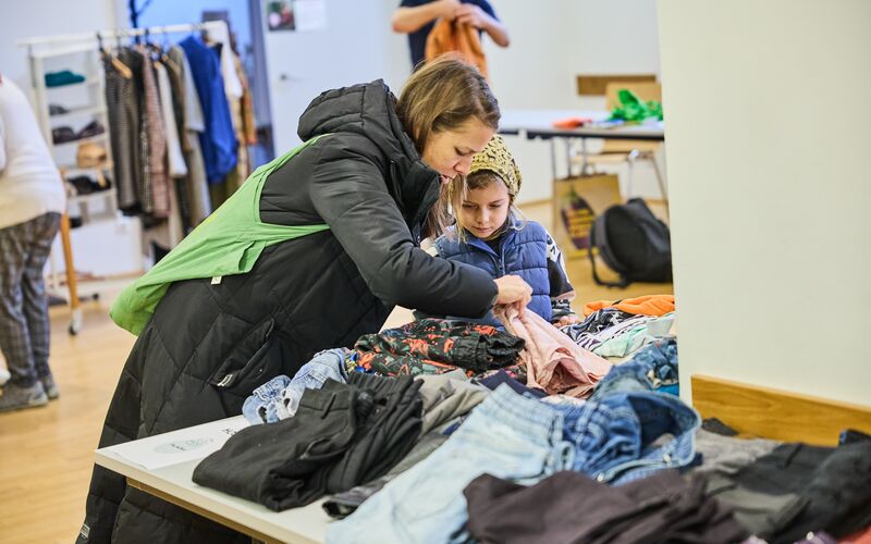 Eine Frau mit einem Kind erkundet einen Tisch voller Second-Hand-Kleidung in einem sozialen Raum.
