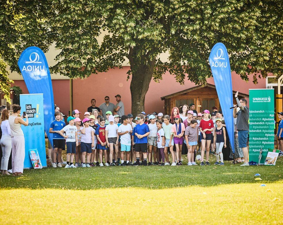 Viele Schüler*innen stehen am Start eines LaufWunders. Es sind Banner und Roll-Ups von Sponsor*innen zu sehen. Ein Mann moderiert die Veranstaltung.