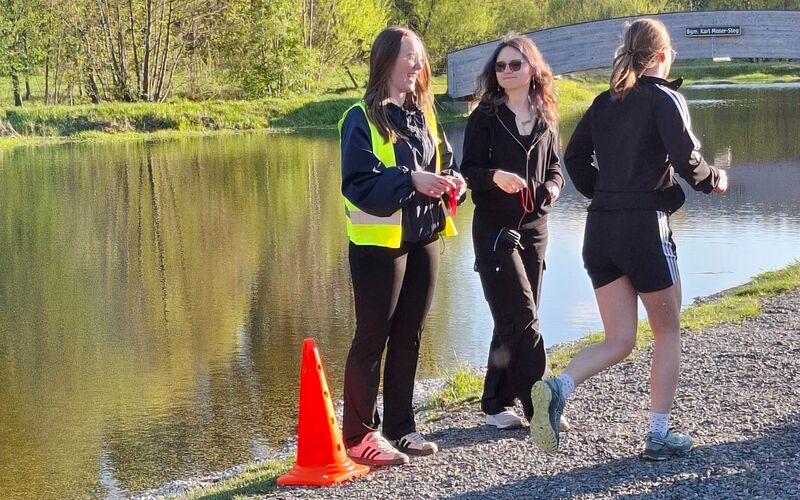 Es sind drei junge Frauen zu sehen. Eine läuft auf einem Schotterweg, die beiden anderen unterhalten sich. Neben ihnen steht ein oranger Leitkegel. Im Hintergrund ist ein Teich zu sehen.