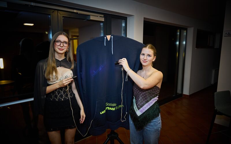 Zwei Frauen präsentieren einen XXL Pullover mit unter UV Licht leuchtenden Fakten zum Thema Fast Fashion