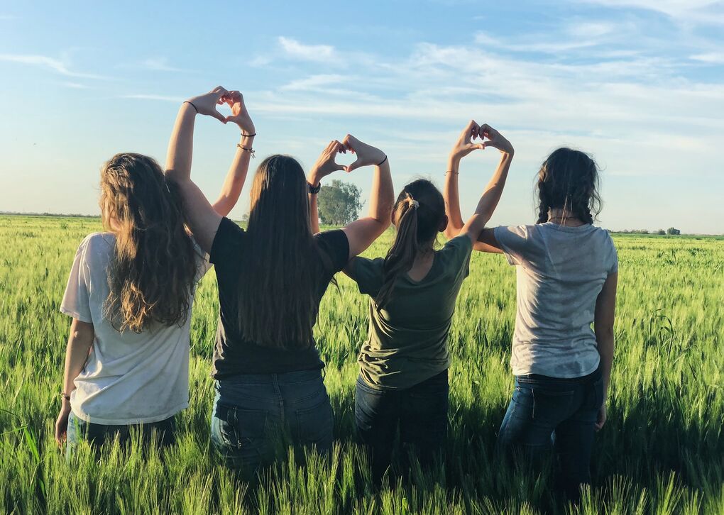 Fünf Frauen stehen in einem Feld und formen mit ihren Händen Herzen. Die Sonne scheint und der Himmel ist blau.