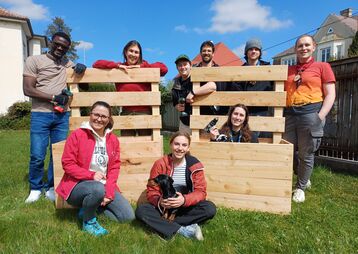Eine Gruppe von Freiwilligen lächelt vor Holzpaletten im Freien, während sie an einem Projekt arbeiten.