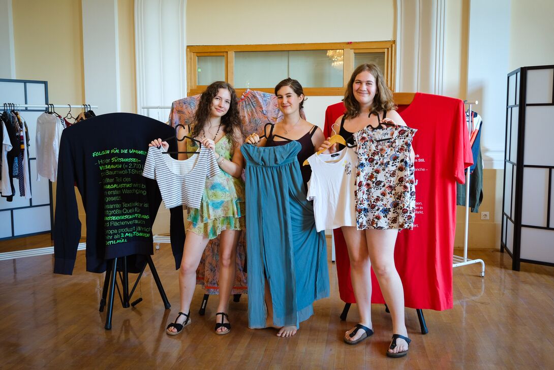 Drei junge Frauen posieren mit Kleidungsstücken in der Hand vor der Ausstellung "tragbar?"