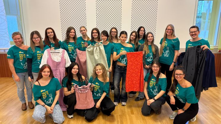 Eine Gruppe von Frauen steht lächelnd zusammen und hält verschiedene Kleider in den Händen, während sie bunte T-Shirts tragen.