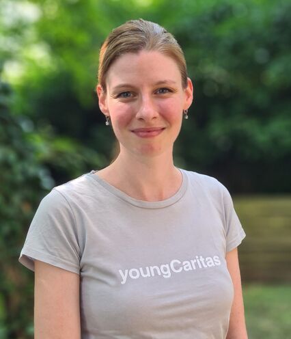 Junge Frau im grauen T-Shirt mit youngCaritas Schriftzug steht vor grünem Hintergrund.