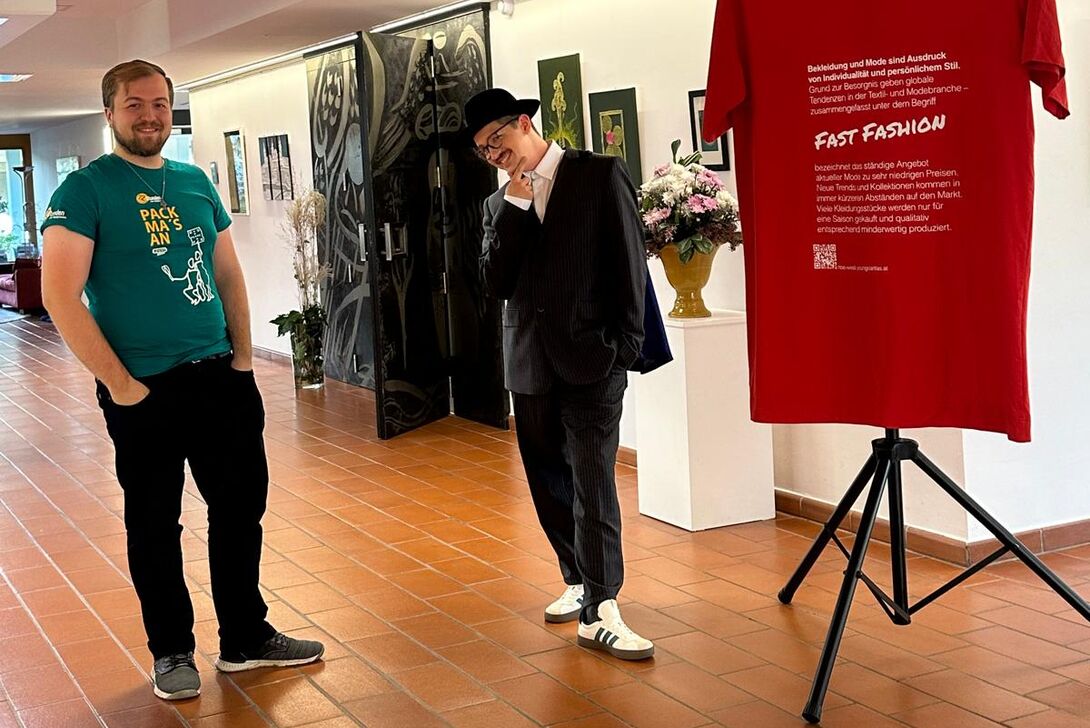 Zwei junge Männer posieren neben einem großen roten T-Shirt, das auf das Thema Fast Fashion aufmerksam macht.