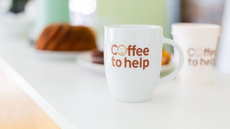 Eine weiße Kaffeetasse mit der Aufschrift 'Coffee to help' steht auf einem Tisch, umgeben von Kuchen und Keksen.