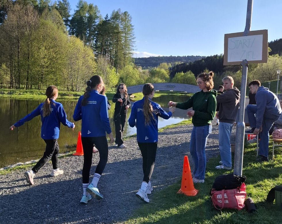Zwei Sportlerinnen übergeben einen Staffelwechsel an einem Gewässer mit Bäumen im Hintergrund während eines Wettkampfs.