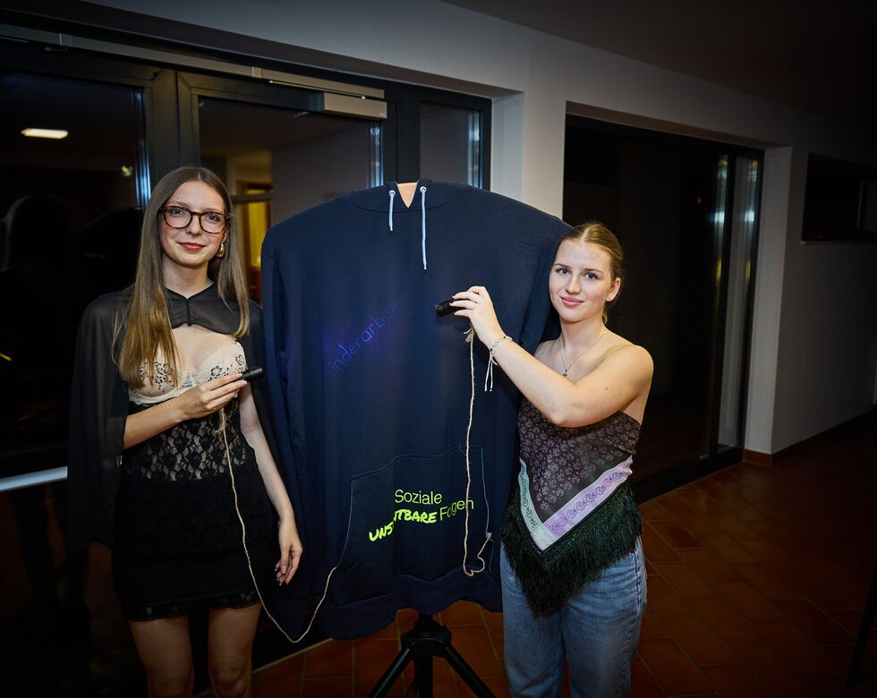 Zwei Frauen präsentieren einen Pullover mit verschiedenen Aufschriften während einer Veranstaltung.