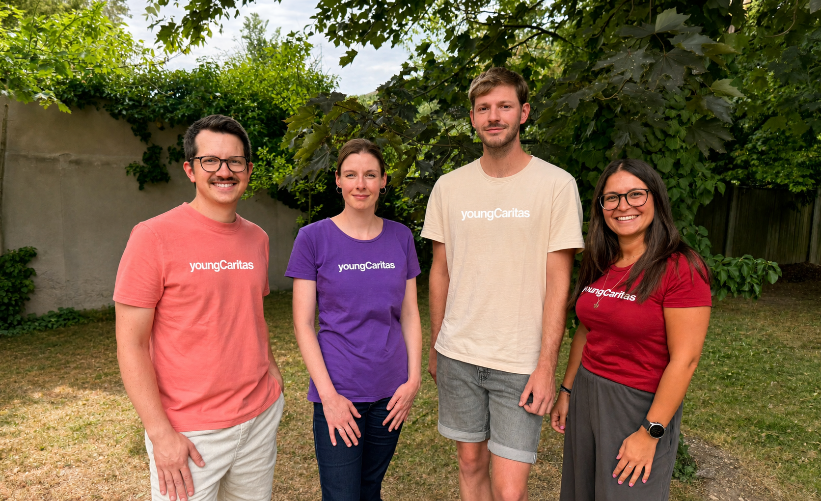 die vier Teammitglieder, zwei Männer in lachsfarbenem und cremfarbenen T-Shirt und zwei Frauen mit violettem und dunkel rotem T-Shirt, alle mit youngCaritas Schriftzug, stehen vor grünem Hintergrund