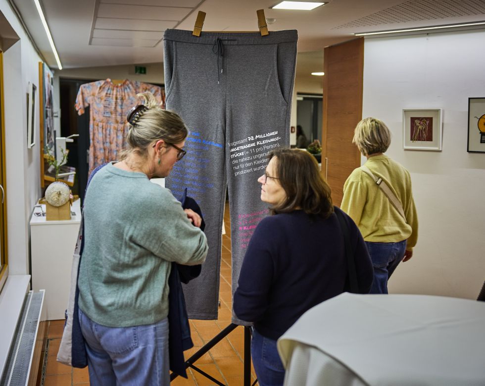 Zwei Frauen diskutieren über eine Kunstinstallation mit grauen Hosen, die Informationen zur Textilproduktion enthalten.
