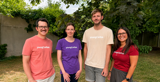 die vier Teammitglieder, zwei Männer in lachsfarbenem und cremfarbenen T-Shirt und zwei Frauen mit violettem und dunkel rotem T-Shirt, alle mit youngCaritas Schriftzug, stehen vor grünem Hintergrund