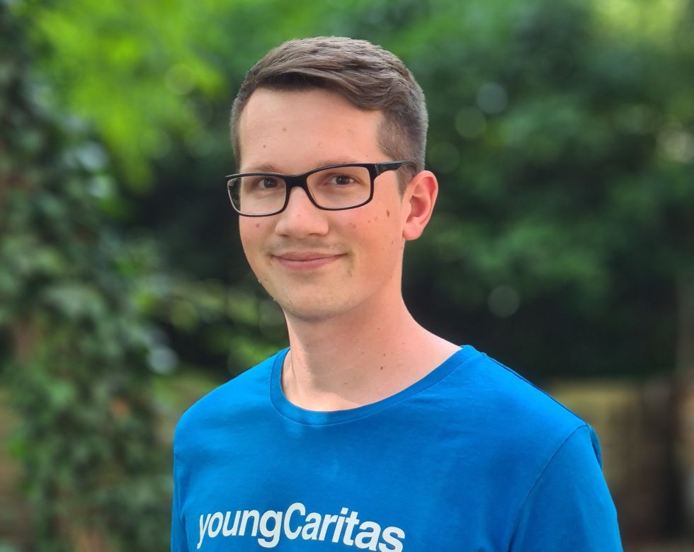 Ein junger Mann trägt ein blaues T-Shirt mit der Aufschrift youngCaritas und lächelt in eine grüne Umgebung.