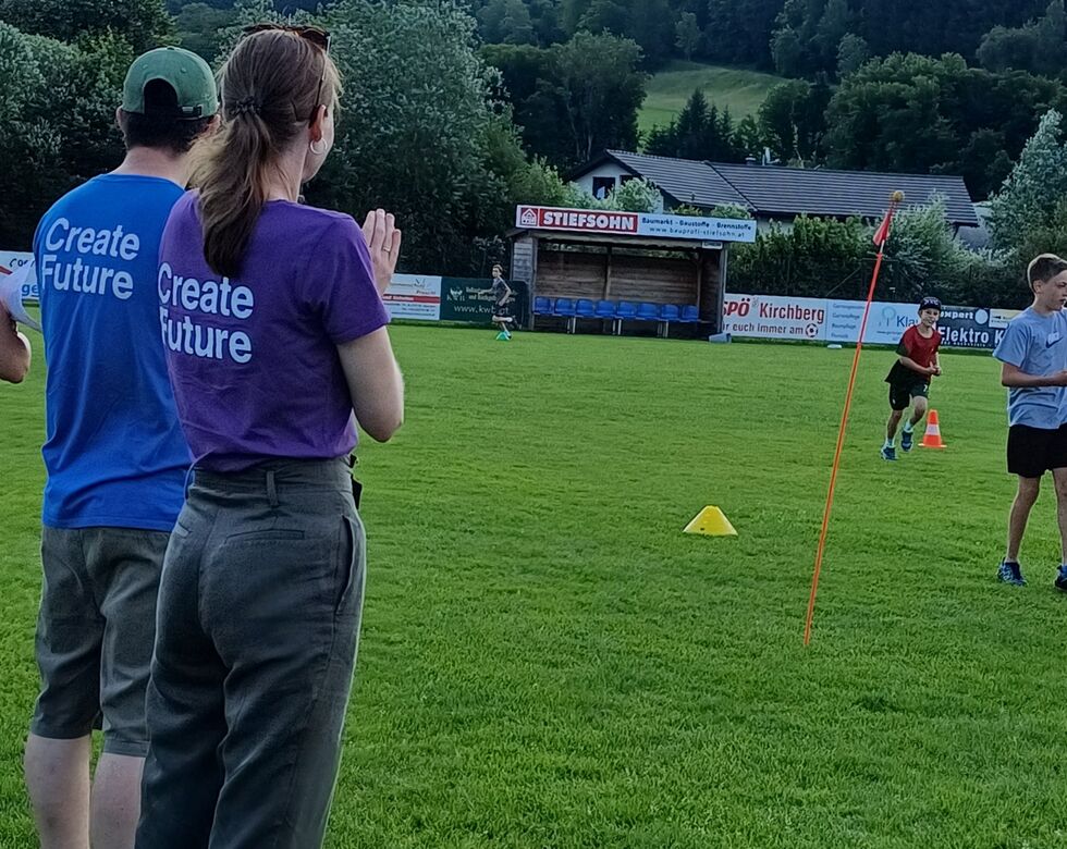 Zwei Personen in T-Shirts mit "Create Future" beobachten ein sportliches Training auf einem grünen Feld, während Kinder aktiv spielen.