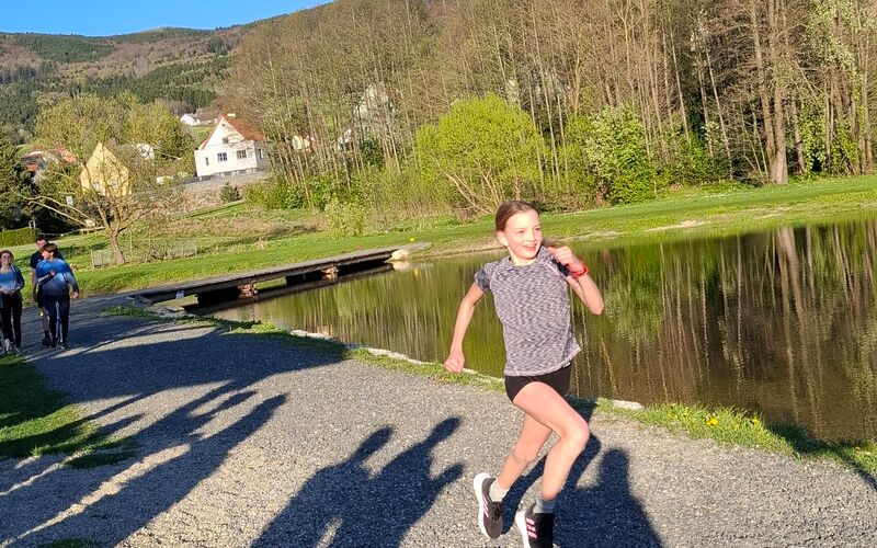 Ein Mädchen läuft auf einem geschotterten Weg. Es sind Schatten von Zuseher*innen am Weg zu sehen.