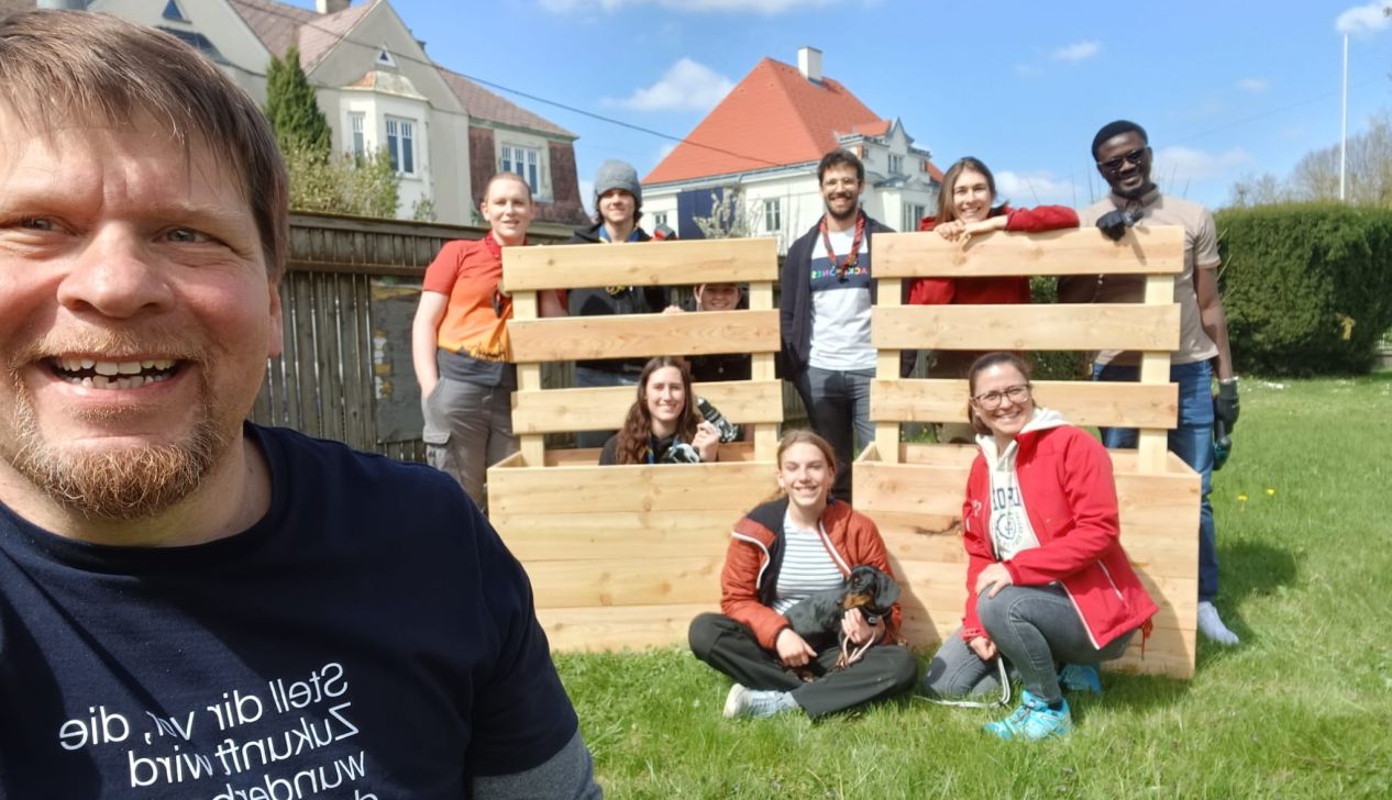 Eine Gruppe von Menschen posiert mit Holzpaletten im Freien, umgeben von grünem Gras und einem klaren blauen Himmel.